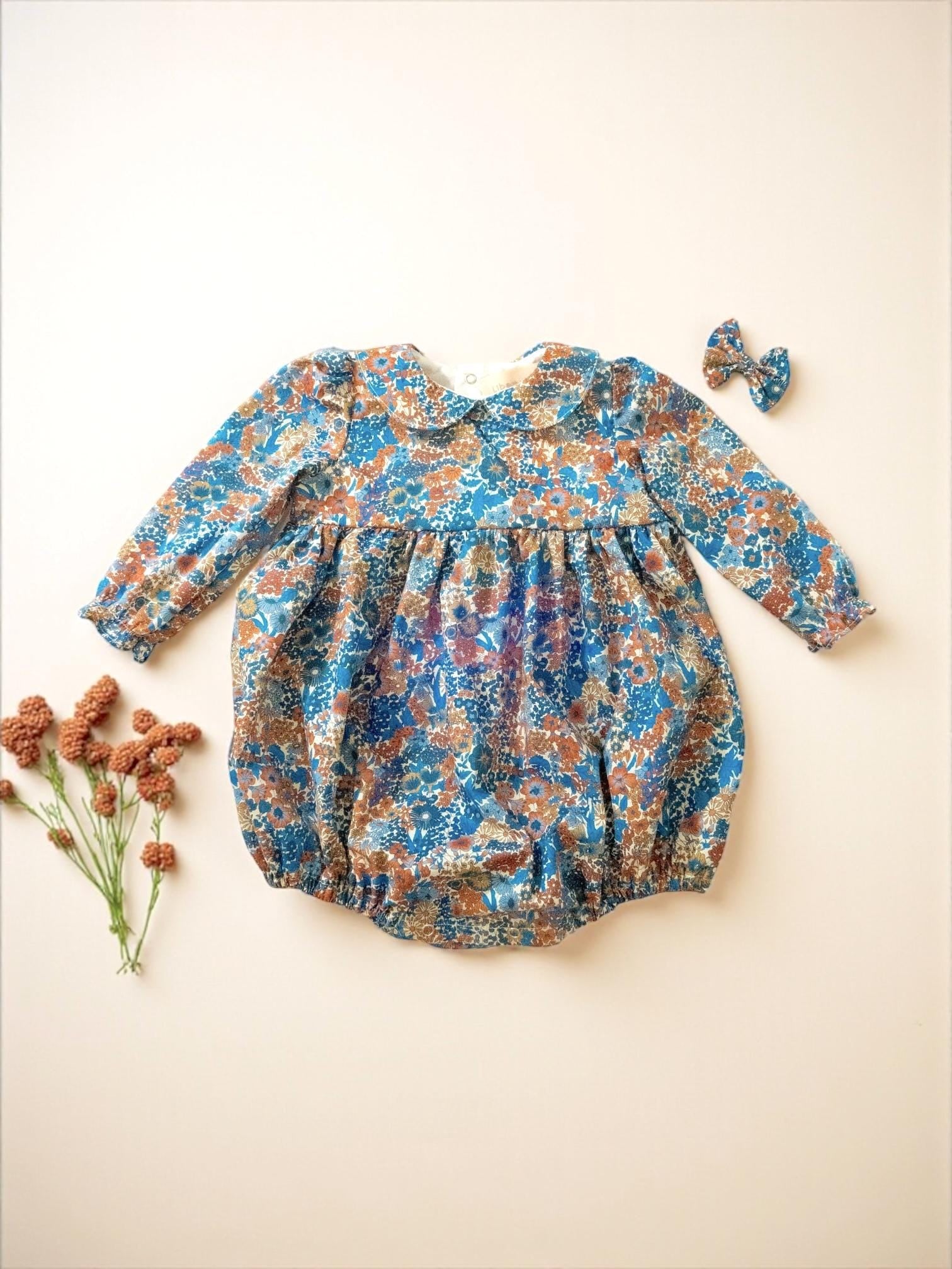 May include: A blue, brown, and white floral print baby romper with long sleeves and a Peter Pan collar. A matching bow hair clip is included. The romper is made of a soft fabric and is designed for comfort.
