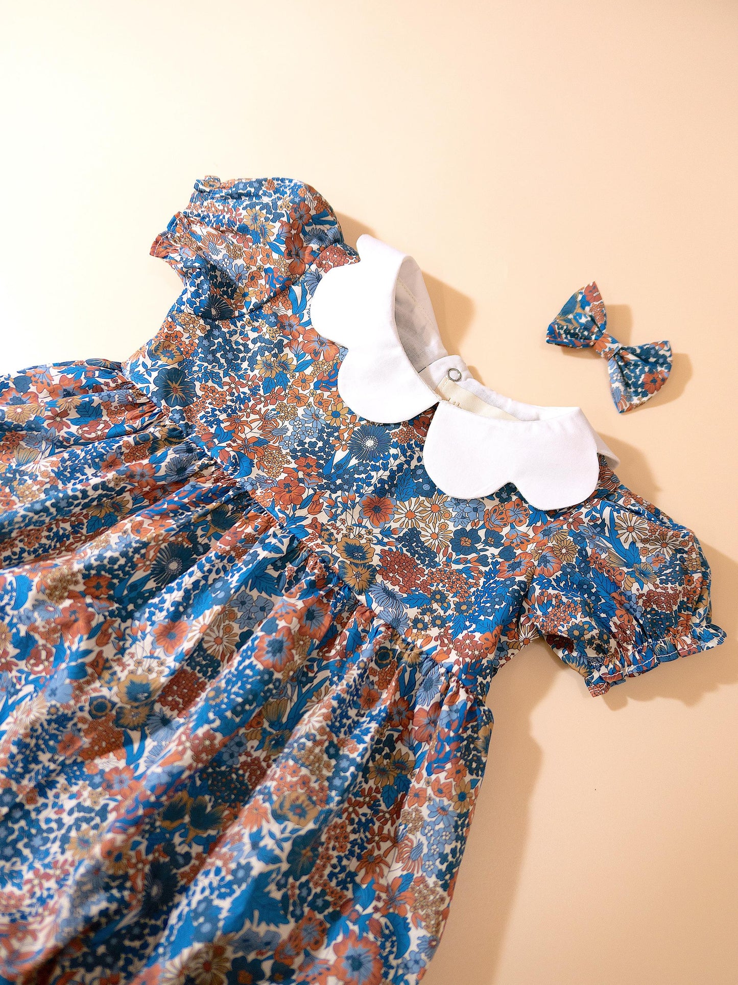 May include: A blue floral print dress with a white Peter Pan collar and matching bow. The dress features short, puffed sleeves and a gathered waist. The floral pattern includes shades of blue, orange, and brown. The dress is designed for a child.