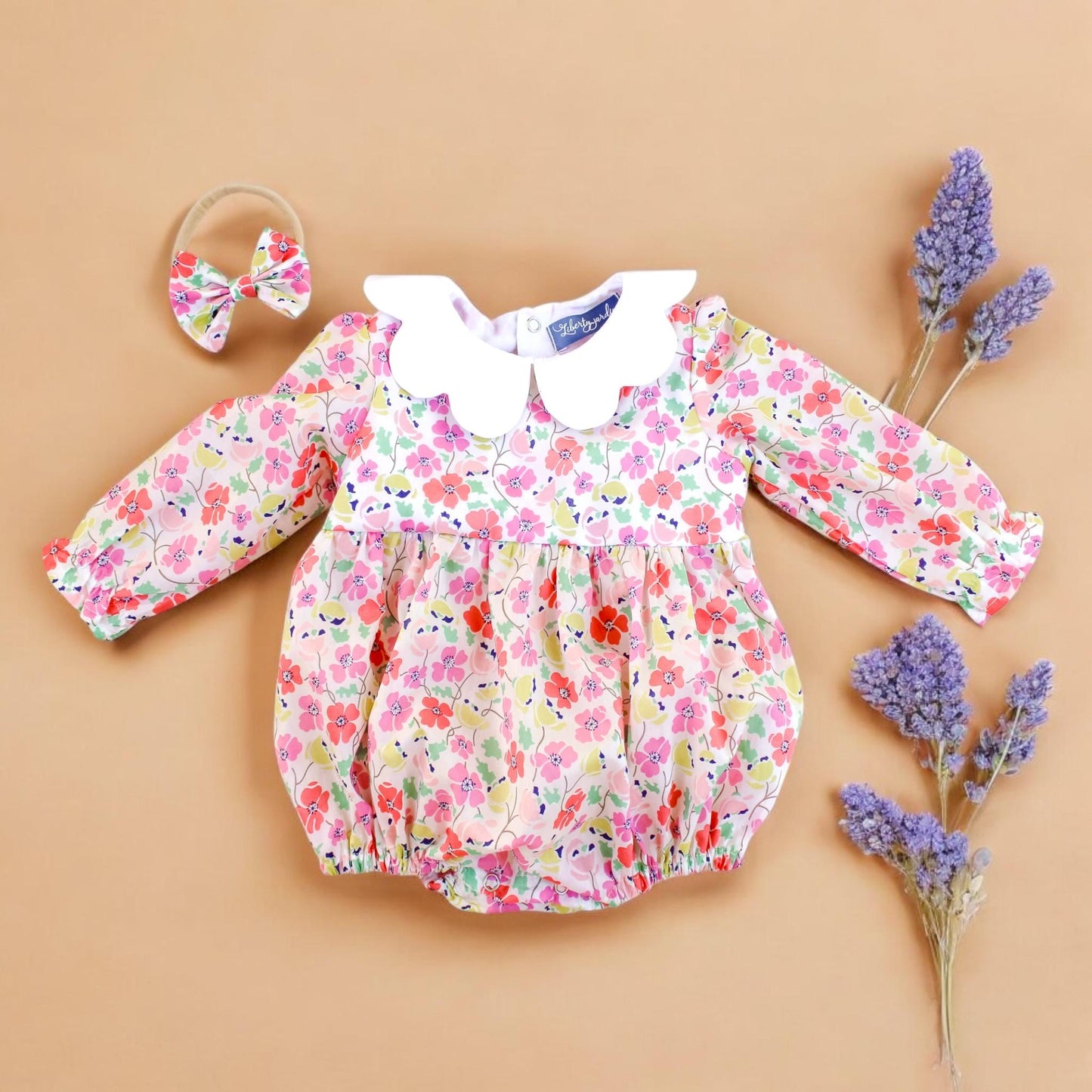 May include: A white baby onesie with a floral print and a white Peter Pan collar. The onesie has long sleeves and a ruffled bottom. A white headband with a bow is included.