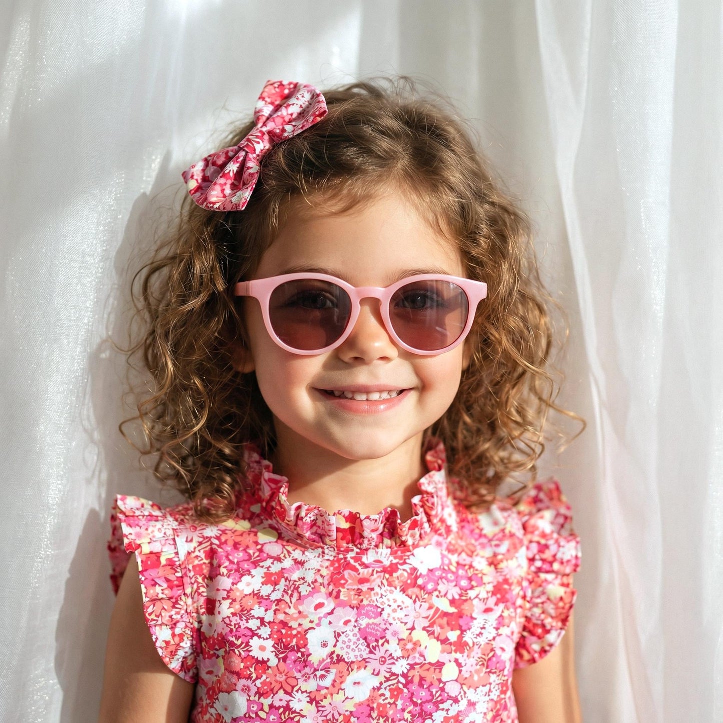 May include: A young girl with curly brown hair wearing pink sunglasses and a matching floral bow. She is also wearing a pink floral dress with a ruffled neckline. The background is a white curtain.
