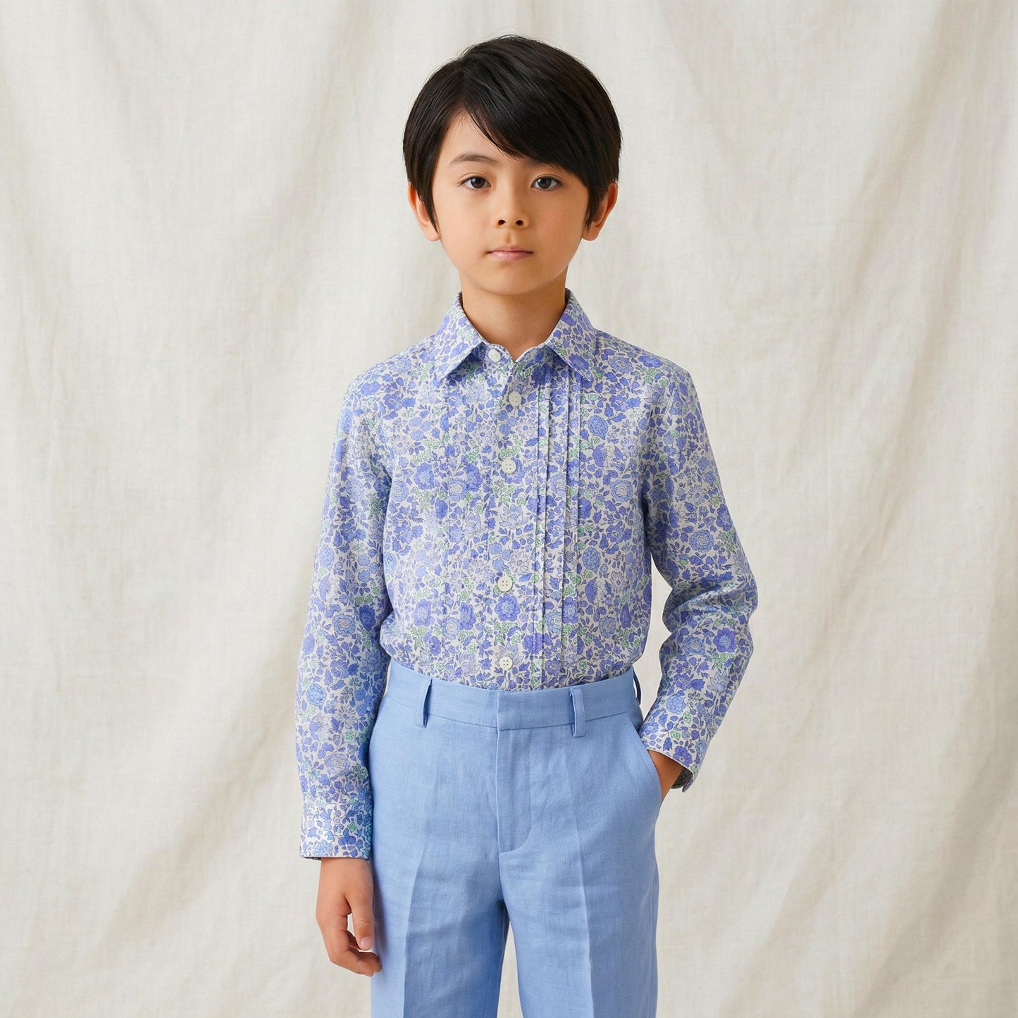 May include: A young person wearing a light blue floral print button-down shirt and matching light blue pants. The shirt has a pleated front and long sleeves. The outfit is suitable for a formal occasion.