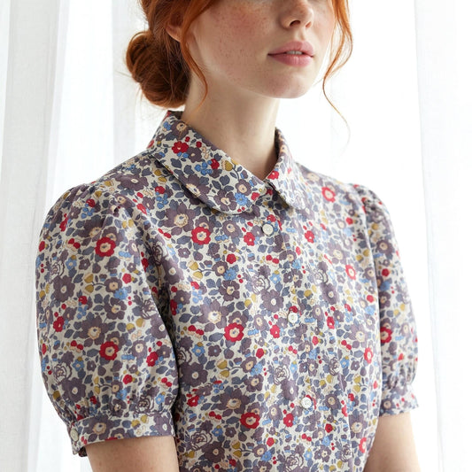 May include: A floral print blouse with a rounded collar and short, puffed sleeves. The fabric features a pattern of small red, blue, and yellow flowers on a muted purple background. The blouse is buttoned up the front.