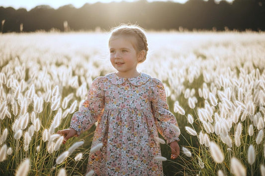 May include: A young child wearing a floral print dress with a Peter Pan collar stands in a field of tall, white, feathery plants. The dress has long sleeves and a pink and yellow floral pattern. The background is blurred with sunlight.