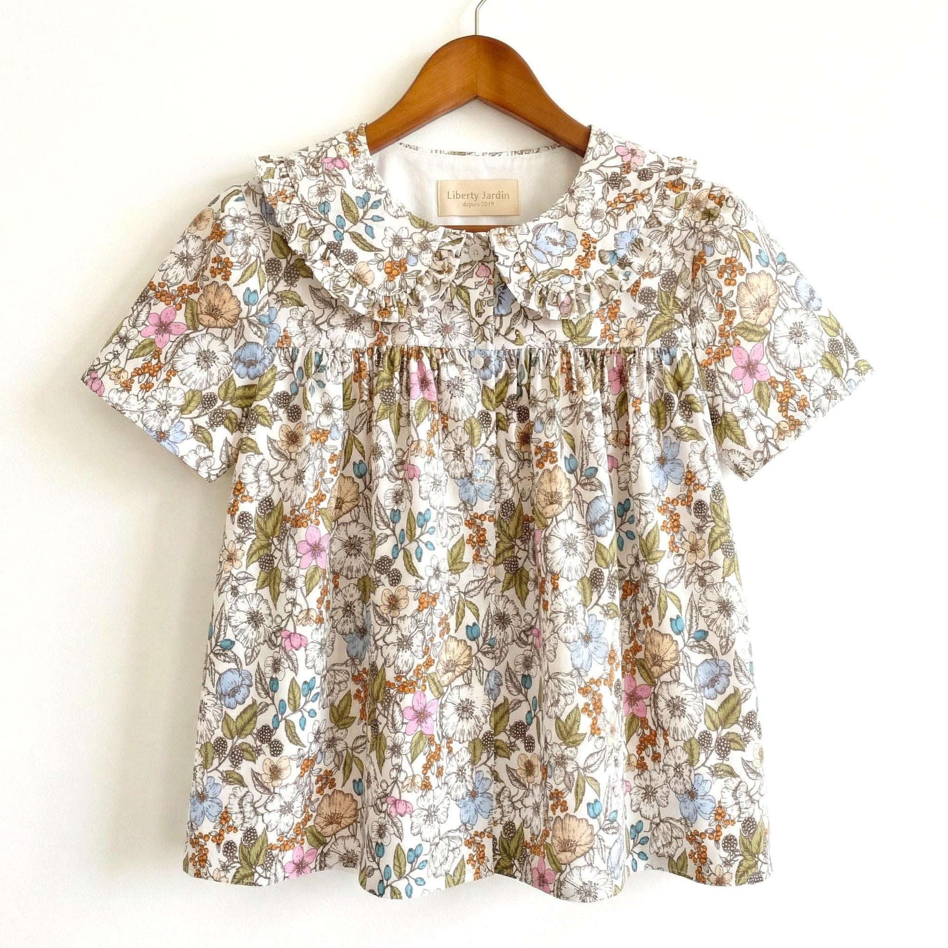 May include: A short-sleeved floral top with a Peter Pan collar. The fabric features a pattern of small flowers in shades of pink, blue, and tan on a white background. The top is hanging on a wooden hanger.