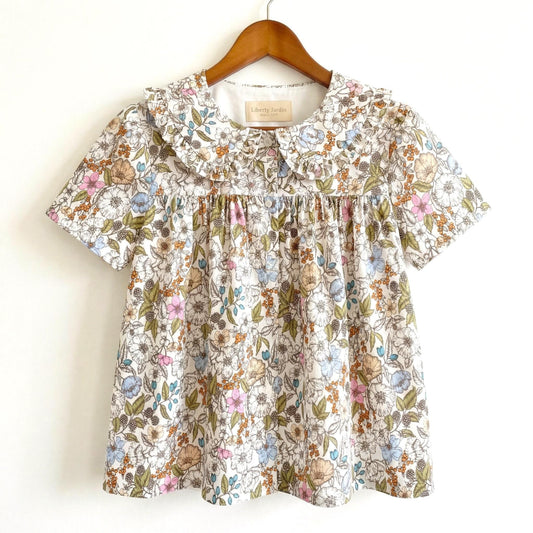 May include: A short-sleeved floral top with a Peter Pan collar. The fabric features a pattern of small flowers in shades of pink, blue, and tan on a white background. The top is hanging on a wooden hanger.