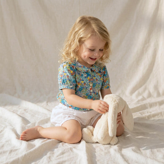 May include: A young child wearing a blue floral print top and white shorts, holding a cream-colored stuffed bunny. The top has a Peter Pan collar and short sleeves. The child is sitting on a white sheet.