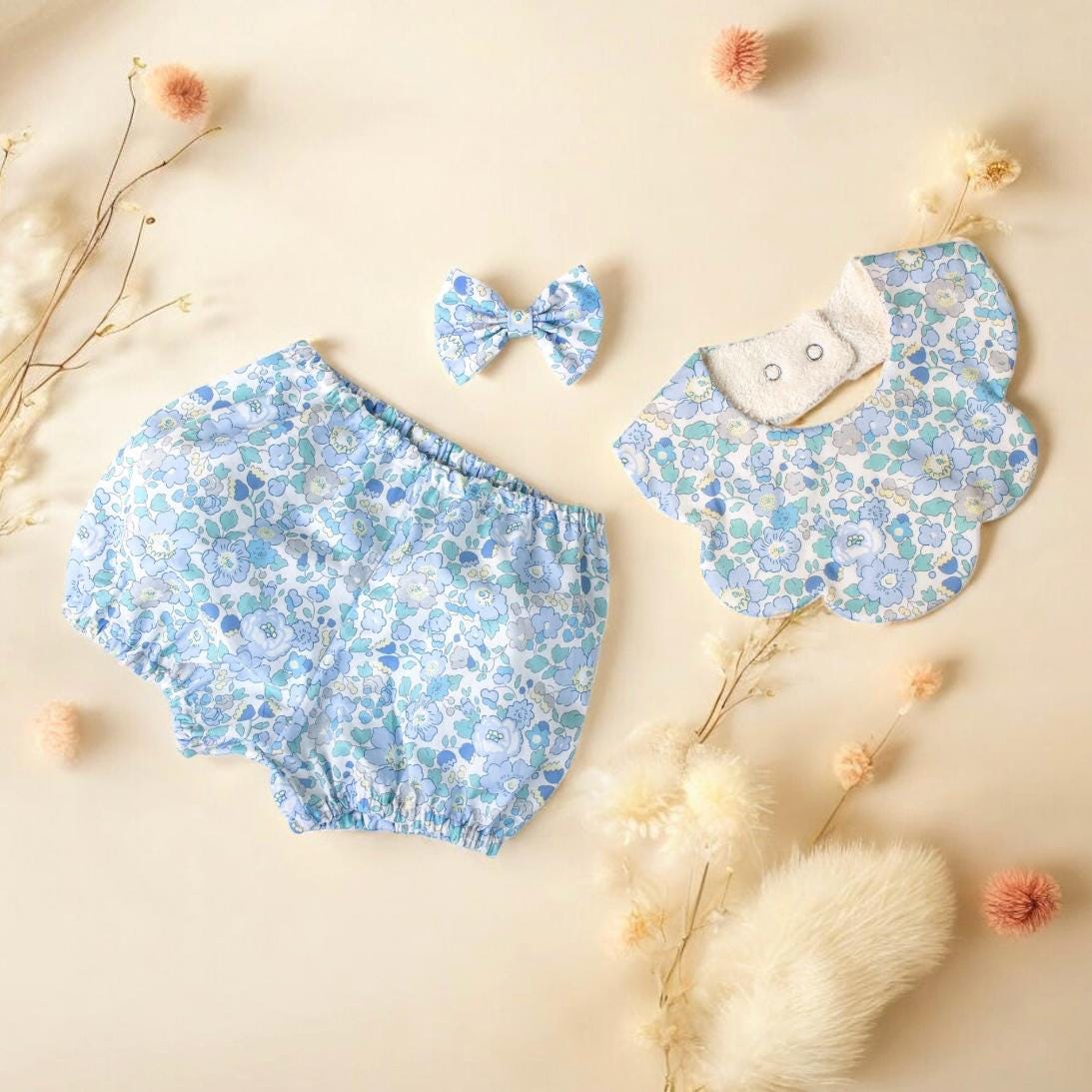 May include: A baby clothing set featuring a pair of bloomers, a bib, and a bow. The items are light blue with a floral pattern in shades of blue and white. The bib has a scalloped edge and two snap closures.