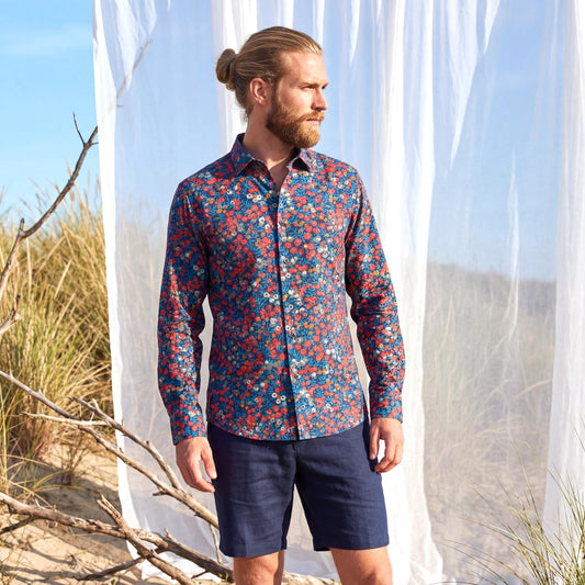 May include: A long-sleeved button-down shirt with a floral pattern in red, white, and yellow on a blue background. The shirt is paired with navy blue shorts. The image is set against a backdrop of tall grass and a white curtain.