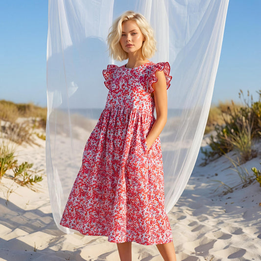 May include: A red floral print dress with ruffled sleeves and pockets. The dress is knee-length and has a loose, flowing silhouette. The background includes a beach setting with sand, plants, and a sheer white curtain.