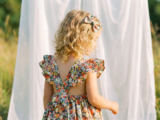 May include: A young child wearing a floral print dress with a bow in their hair. The dress features a V-back and ruffled sleeves. The floral pattern includes red, yellow, and blue flowers on a dark background.