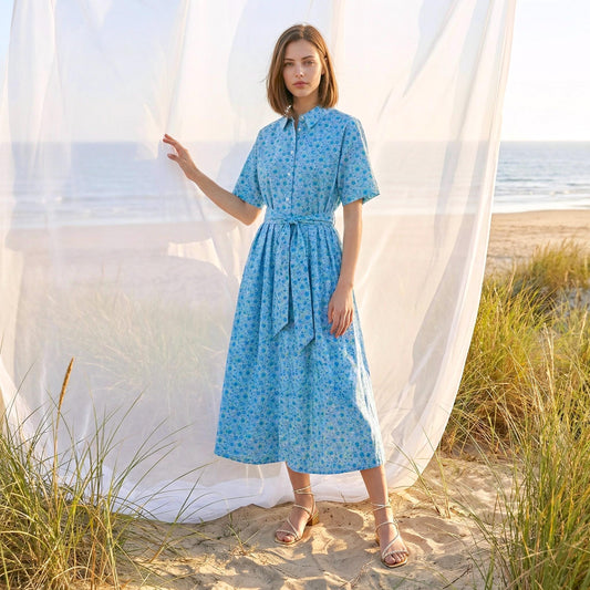 May include: A light blue floral print midi dress with short sleeves and a matching belt. The dress features a button-down front and is worn with strappy sandals. The background includes a beach scene with sand, sea, and tall grass.