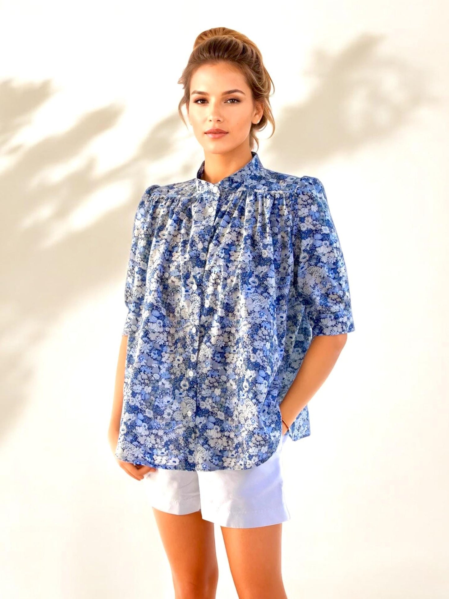 May include: A blue floral print blouse with a mandarin collar and short sleeves. The blouse is paired with white shorts. The floral pattern features shades of blue and white. The model is standing in front of a white background.