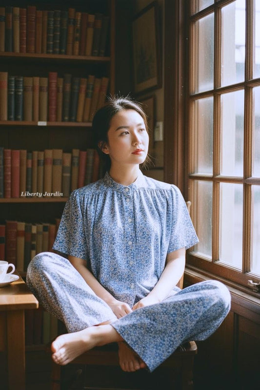 May include: A person wearing a blue floral print pajama set sits by a window. The set includes a button-down shirt and matching pants. The text "Liberty Jardin" is visible on the bookshelf in the background. A cup and saucer sit on a small table.