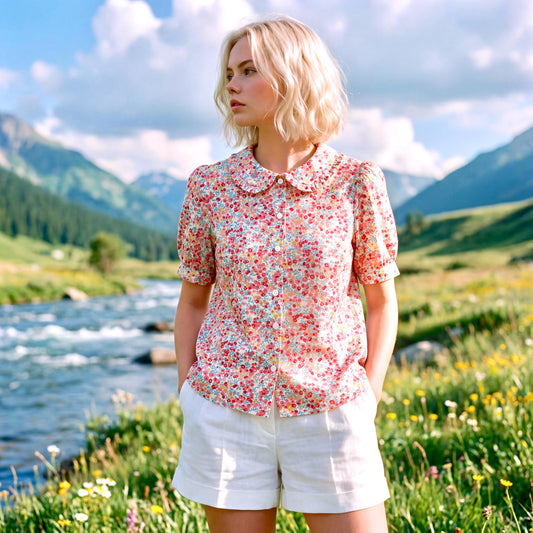 May include: A floral print blouse with a rounded collar and short sleeves. The blouse is buttoned down and paired with white shorts. The background features a river, mountains, and a blue sky with clouds, creating a scenic outdoor setting.