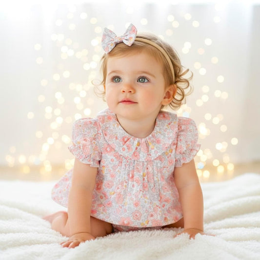 May include: A baby wearing a pink floral print outfit, including a dress and matching headband with a bow. The outfit has a Peter Pan collar and short sleeves. The baby is sitting on a white, fluffy surface.