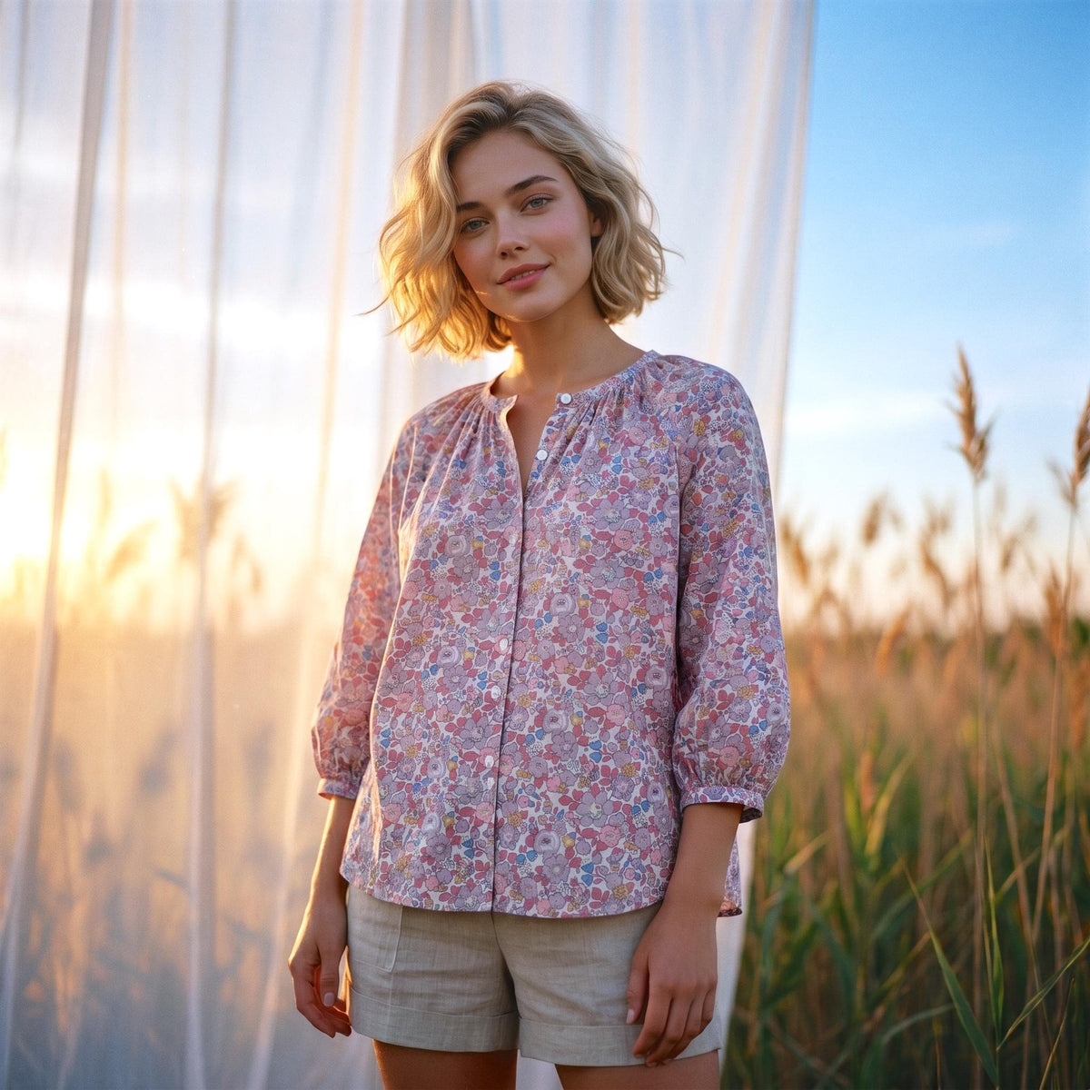 May include: A light pink floral print blouse with three-quarter sleeves and a button-down front. The blouse is paired with beige shorts. The background features tall grass and a sheer white curtain.