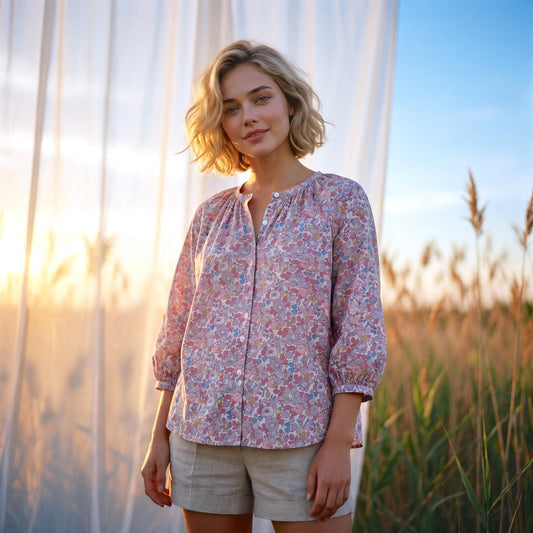 May include: A light pink floral print blouse with three-quarter sleeves and a button-down front. The blouse is paired with beige shorts. The background features tall grass and a sheer white curtain.