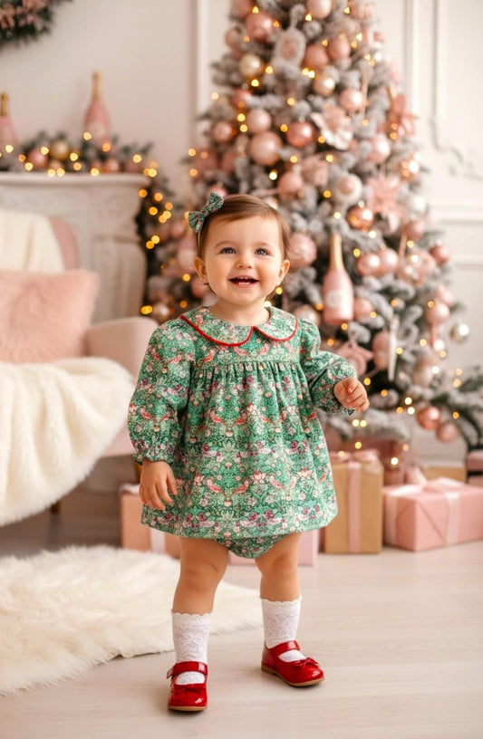 May include: A smiling toddler wearing a green floral print dress with a red-trimmed collar and matching bloomers. The outfit is paired with red Mary Jane shoes and white lace socks. A Christmas tree is in the background.